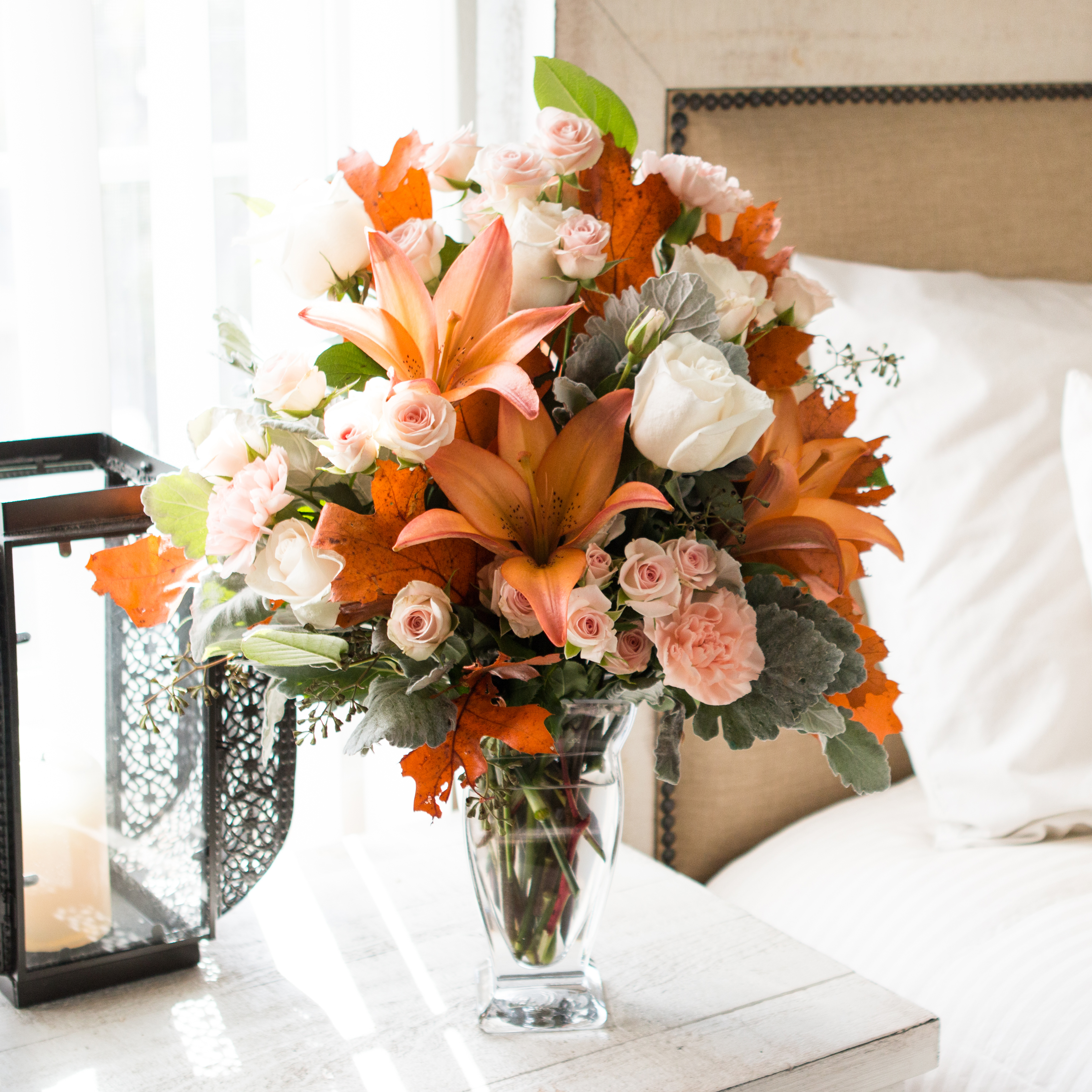 Orange lilies, light pink roses, greenery and more in a clear vase on a bedside table