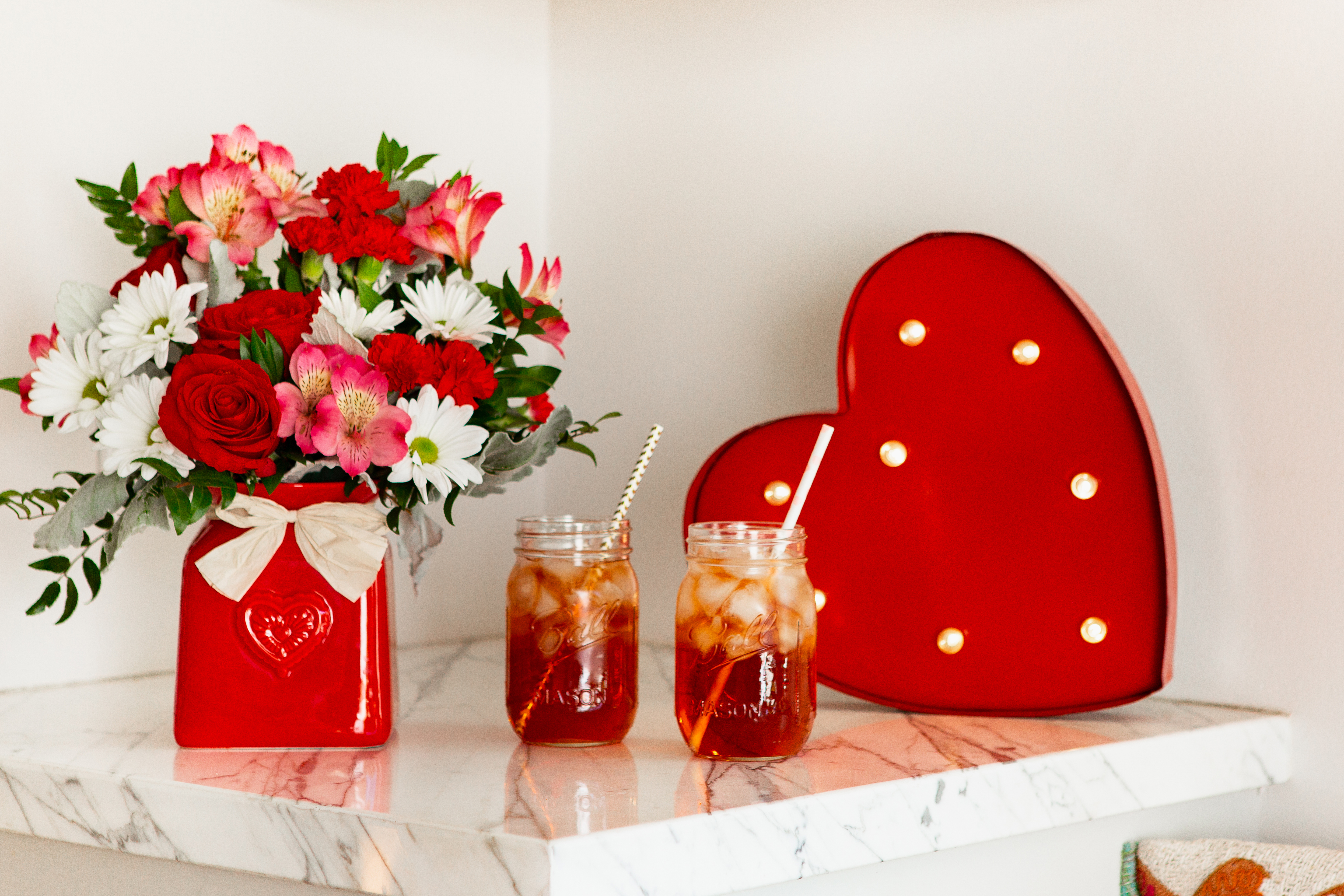 Red roses, white daises, and more in a red vase next to tea glasses and a red heart