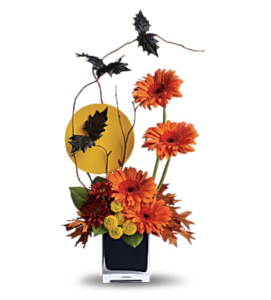 Orange gerberas, yellow mums, and more fill a cube vase with flying bats above