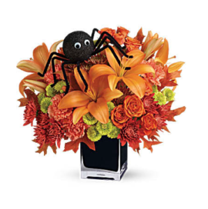 Orange lilies, roses, carnations, and more fill a cube vase with a spider on top of the flowers